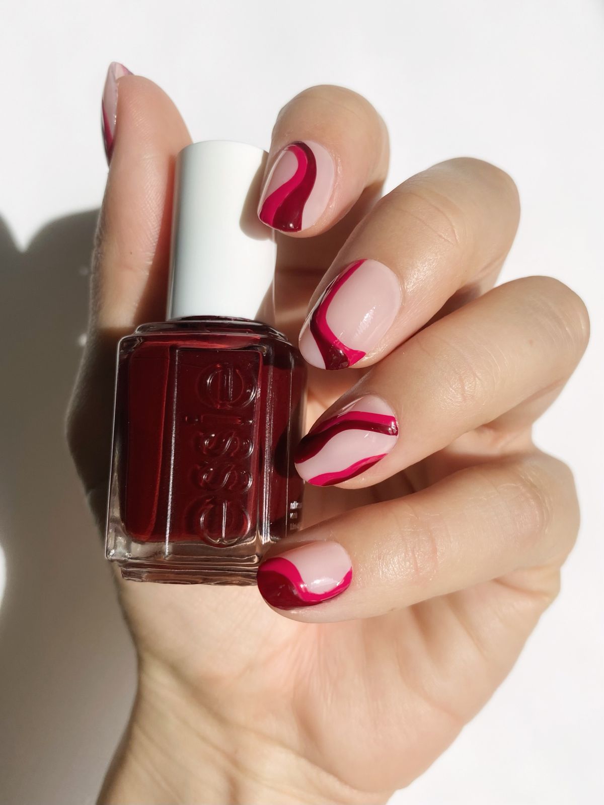 a hand with abstract nail polish designs holding a berry coloured bottle of Essie nail polish