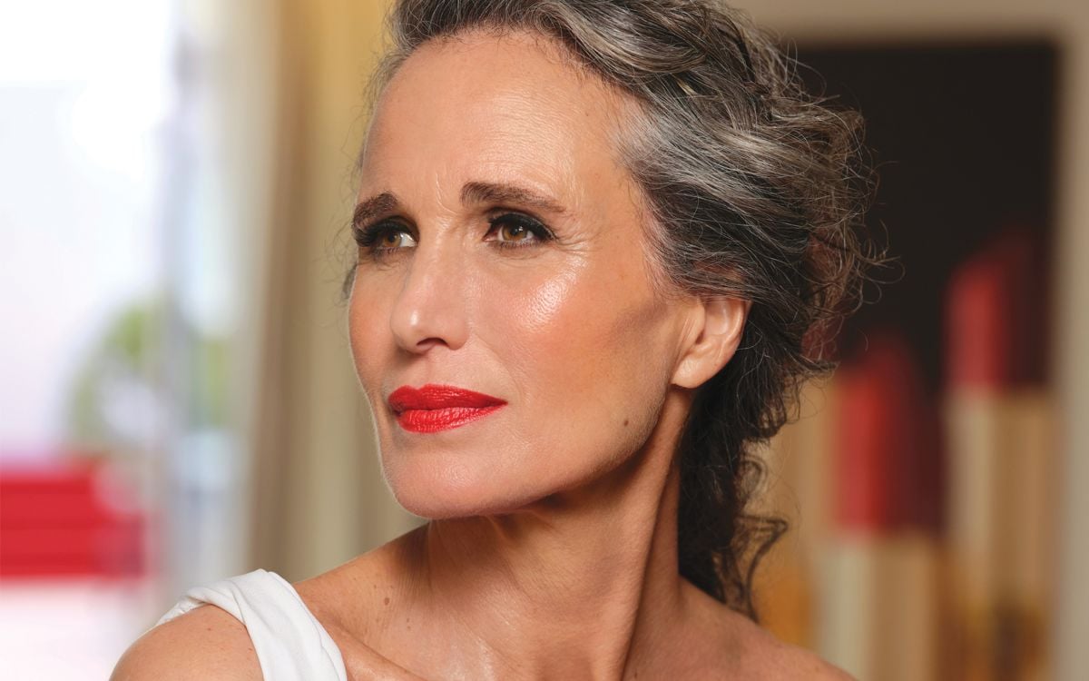 Andie Macdowell close up, wearing red lipstick and other beauty products such as blush and eyeliner. Her hair is back into a loose updo