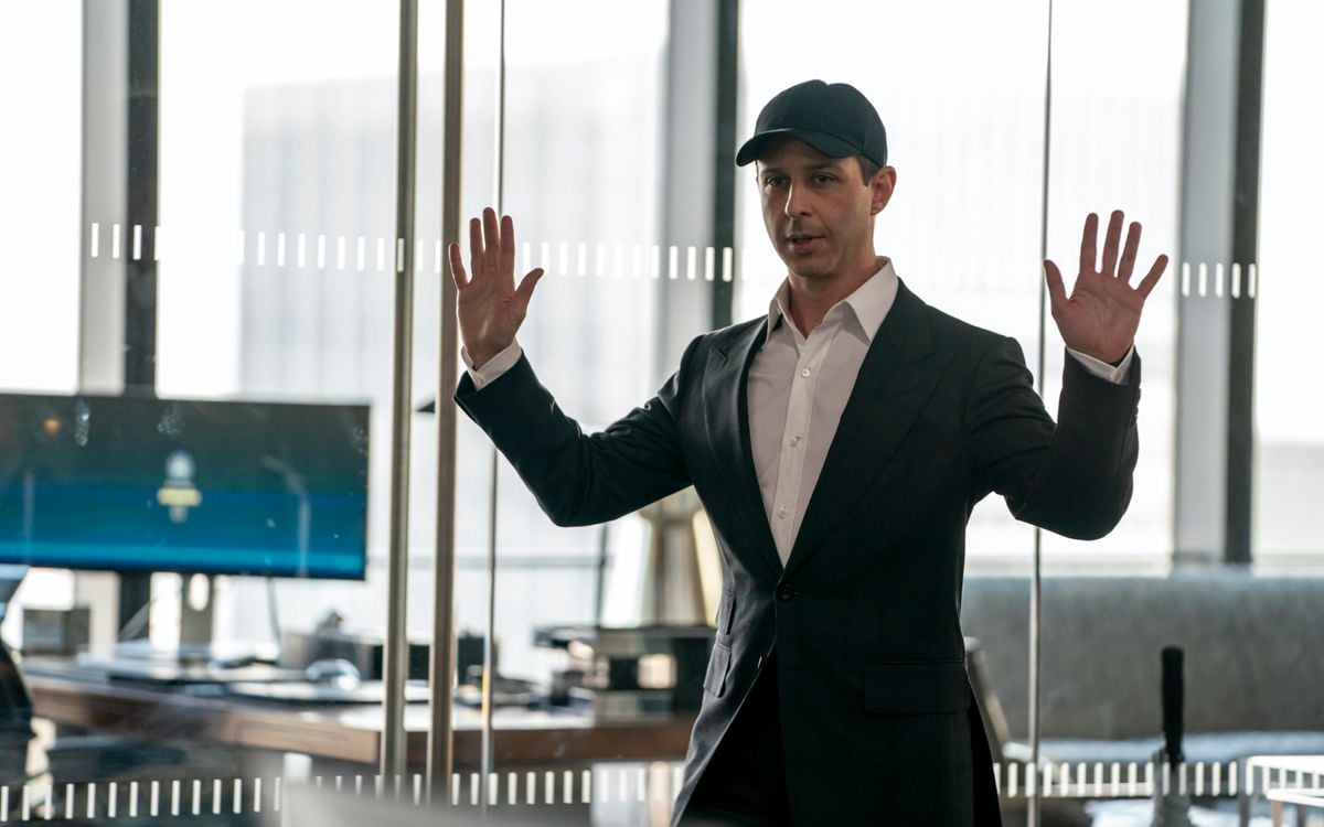 a still from the tv show succession of kendall roy wearing a white dress shirt, dark blazer and black baseball hat