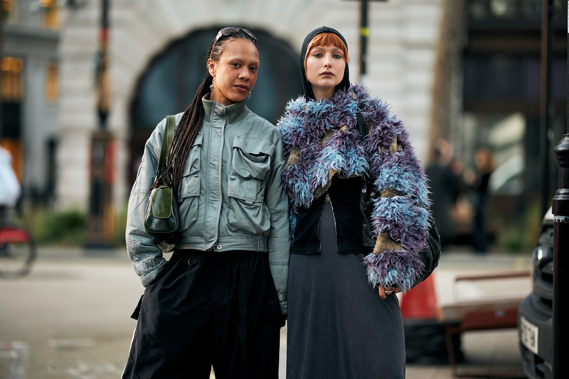 Two people stand on a city street. One wears a grey jacket and black pants, carrying a green bag. The other has a colorful, textured jacket over a black top and grey dress, and a black hat. This image invites reflection on how to find your style even within urban trends, and the question of why are we dressing the same, given the differences in their outfits.