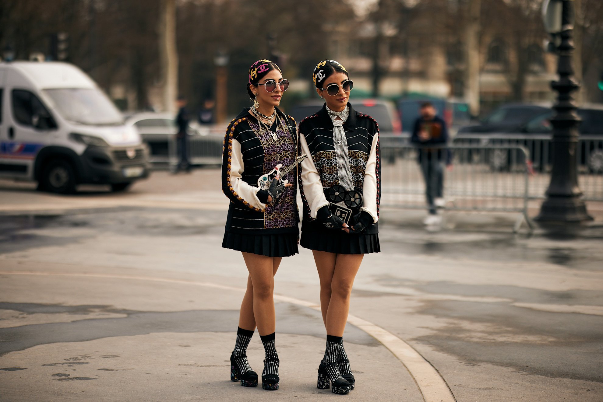 Two individuals with strikingly similar outfits stand on a city street, prompting the question: why are we dressing the same? They wear patterned headbands, embellished sunglasses, layered tops, black pleated skirts, patterned socks, and platform heels, suggesting a shared fashion trend. This image invites viewers to consider how to find your style within or beyond such trends.